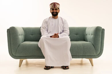 مفهوم الرجولة والثقة بالنفس، الاهتمام بالأناقة والمظهر الخارجي، بورتريه لرجل عربي خليجي عماني يرتدي الدشداشة و العمامة يجلس على الاريكة ينظر الى الكاميرا بتعابير السرور، خلفية بيضاء