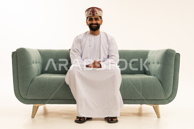 الاهتمام بالأناقة والمظهر الخارجي، مفهوم الرجولة والوقار، ايماءت الثقة بالنفس، بورتريه لرجل عربي خليجي عماني يرتدي الدشداشة و العمامة يجلس على الاريكة ينظر الى الكاميرا بتعابير السرور، خلفية بيضاء