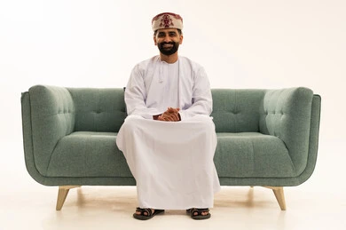 الاهتمام بالأناقة والمظهر الخارجي، مفهوم الرجولة والوقار، ايماءت الثقة بالنفس، بورتريه لرجل عربي خليجي عماني يرتدي الدشداشة و العمامة يجلس على الاريكة ينظر الى الكاميرا بتعابير السرور، خلفية بيضاء