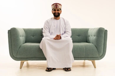 مفهوم الرجولة والوقار، ايماءت الثقة بالنفس، الاهتمام بالأناقة والمظهر الخارجي، بورتريه لرجل عربي خليجي عماني يرتدي الدشداشة و العمامة يجلس على الاريكة ينظر الى الكاميرا بتعابير السرور، خلفية بيضاء