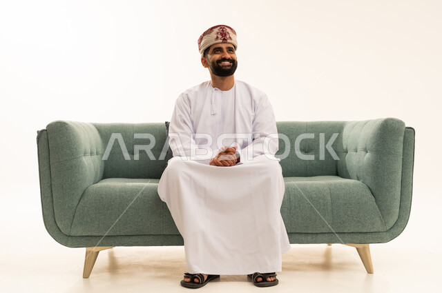 النظر الى شيء ما بايماءات وجه تدل على السعادة و السرور, مفهوم الرجولة والثقة بالنفس، الاهتمام بالأناقة والمظهر الخارجي، بورتريه لرجل عربي خليجي عماني يرتدي الدشداشة و العمامة يجلس على الاريكة، خلفية بيضاء