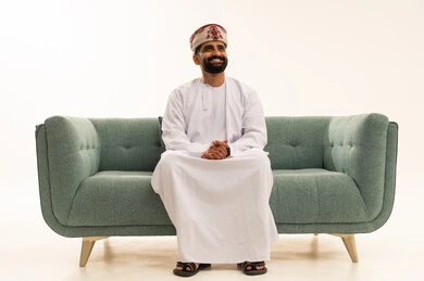 Looking at something with facial expressions that indicate happiness and joy, the concept of masculinity and self-confidence, attention to elegance and appearance, a portrait of an Arab Gulf Omani man wearing a dishdasha and a turban sitting on a sofa, white background.