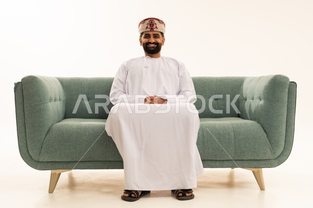 ايماءات الثقة بالنفس، مفهوم الرجولة والوقار، الاهتمام بالأناقة والمظهر الخارجي، بورتريه لرجل عربي خليجي عماني يرتدي الدشداشة و العمامة يجلس على الاريكة ينظر الى الكاميرا بتعابير السرور، خلفية بيضاء