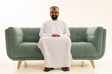 Gestures of self-confidence, the concept of masculinity and dignity, attention to elegance and appearance, a portrait of an Arab Gulf Omani man wearing a dishdasha and a turban sitting on a sofa looking at the camera with expressions of joy, white background.