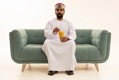 انشطة ترفيهية منزلية، مشاهدة الافلام والعروض المسرحية،  بورتريه لرجل عربي خليجي عماني يرتدي الدشداشة و العمامة يجلس على الاريكة يحمل بيده دلو الفشار، النظر الى الكاميرا بتعابير الاندهاش والاستغراب، تناول الوجبات الخفيفة والسناك، خلفية بيضاء