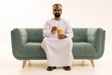 مفهوم التسليه والترفيه، قضاء أوقات ممتعة في عطلة نهاية الاسبوع، أنشطة وفعاليات مسلية، الاستعداد لمشاهدة فيلم في السينما، بورتريه لرجل عربي خليجي عماني يرتدي الدشداشة و العمامة يجلس على الاريكة يحمل بيده علبة من الفشار المقرمش، خلفية بيضاء