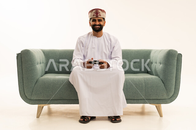 تعابير الاستمتاع باللعب بالألعاب الالكترونية الترفيهية، استخدام الأجهزة التكنولوجية والتقنيات الحديثة، بورتريه لرجل عربي خليجي عماني يرتدي الدشداشة و العمامة يجلس على الاريكة يمسك بيده عصا التحكم بإيماءات السرور والاندماج، خلفية بيضاء