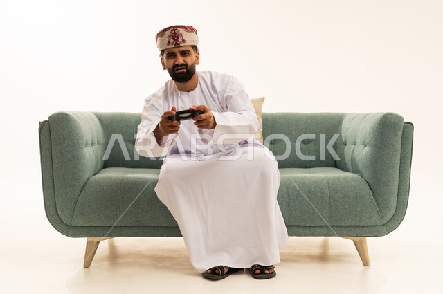 استخدام الأجهزة التكنولوجية والتقنيات الحديثة، تعابير الاستمتاع باللعب بالألعاب الالكترونية الترفيهية، بورتريه لرجل عربي خليجي عماني يرتدي الدشداشة و العمامة يجلس على الاريكة يمسك بيده عصا التحكم بإيماءات السرور والاندماج، خلفية بيضاء