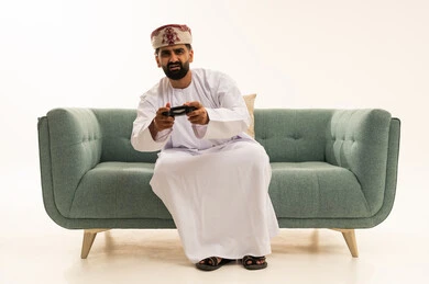 استخدام الأجهزة التكنولوجية والتقنيات الحديثة، تعابير الاستمتاع باللعب بالألعاب الالكترونية الترفيهية، بورتريه لرجل عربي خليجي عماني يرتدي الدشداشة و العمامة يجلس على الاريكة يمسك بيده عصا التحكم بإيماءات السرور والاندماج، خلفية بيضاء