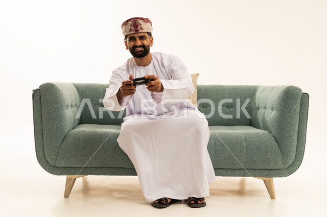 Expressions of enjoyment while playing recreational electronic games, using technological devices and modern techniques, a portrait of an Arab Gulf Omani man wearing a dishdasha and a turban sitting on a sofa holding a game controller with gestures of joy and immersion, white background.