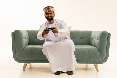 تعابير الاستمتاع باللعب بالألعاب الالكترونية الترفيهية، استخدام الأجهزة التكنولوجية والتقنيات الحديثة، بورتريه لرجل عربي خليجي عماني يرتدي الدشداشة و العمامة يجلس على الاريكة يمسك بيده عصا التحكم بإيماءات السرور والاندماج، خلفية بيضاء