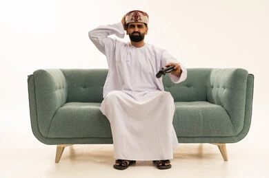 تقنيات الكترونية حديثة, إيماءات الحزن والخسارة, قضاء أوقات الفراغ باللعب في ألعاب البلايستيشن الترفيهية،  بورتريه لرجل عربي خليجي عماني يرتدي الدشداشة و العمامة يجلس على الاريكة يحمل عصا التحكم يضع يدية على رأسه، خلفية بيضاء