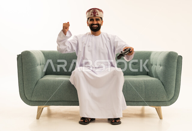 ايماءات الانتصار والفوز، التفاعل والاندماج مع ألعاب البلايستيشن، تقنيات الكترونية ترفيهيه, بورتريه لرجل عربي خليجي عماني يرتدي الدشداشة و العمامة يجلس على الاريكة يحمل بيده عصا التحكم، تجربة ترفيهية ممتعة ومثيرة في الأجهزة الحديثة، خلفية بيضاء