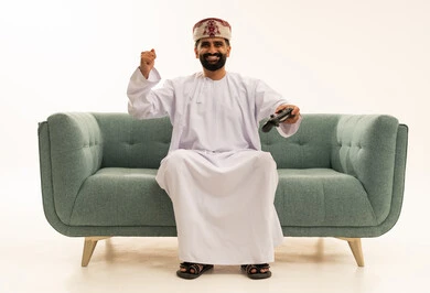 ايماءات الانتصار والفوز، التفاعل والاندماج مع ألعاب البلايستيشن، تقنيات الكترونية ترفيهيه, بورتريه لرجل عربي خليجي عماني يرتدي الدشداشة و العمامة يجلس على الاريكة يحمل بيده عصا التحكم، تجربة ترفيهية ممتعة ومثيرة في الأجهزة الحديثة، خلفية بيضاء