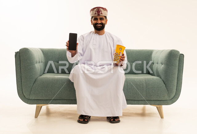 عرض خلفية شاشة سوداء على الهاتف المحمول، تناول الفوشار اللذيذ،بورتريه لرجل عربي خليجي عماني يرتدي الدشداشة و العمامة يجلس على الاريكة يحمل بيده علبة من الفشار الطازة ، الترفيه عن النفس وقضاء وقت ممتع، خلفية بيضاء
