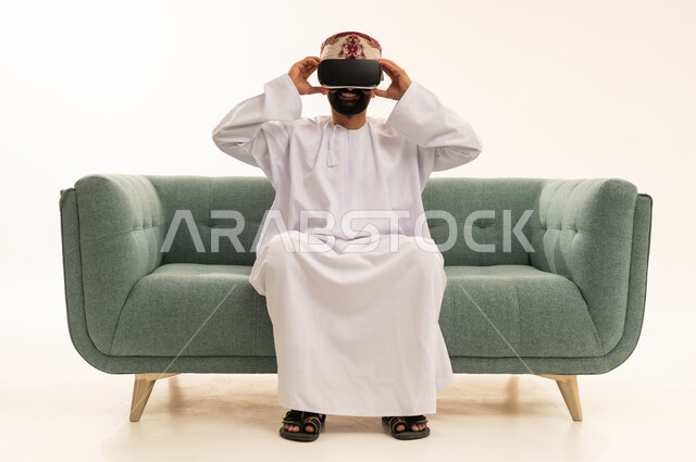 الاندماج ومحاكاة العالم الخيالي، المتعة والتسلية بالمشاهدة عبر نظارة الواقع الافتراضي، بورتريه لرجل عربي خليجي عماني يرتدي الدشداشة و العمامة يجلس على الاريكة  يضع نظارات ثلاثية الأبعاد (الفي ار), التطور التقني والتكنولوجي، خلفية بيضاء