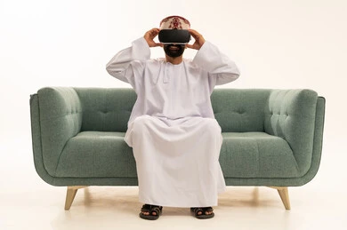الاندماج ومحاكاة العالم الخيالي، المتعة والتسلية بالمشاهدة عبر نظارة الواقع الافتراضي، بورتريه لرجل عربي خليجي عماني يرتدي الدشداشة و العمامة يجلس على الاريكة  يضع نظارات ثلاثية الأبعاد (الفي ار), التطور التقني والتكنولوجي، خلفية بيضاء