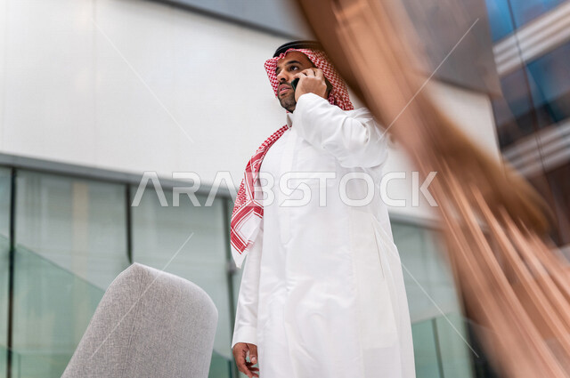 رجل خليجي سعودي ، يقوم بإجراء مكالمة هاتفية عبر الهاتف المحمول في داخل الفندق السياحي ، إستخدام أجهزة تقنية حديثة ، جلسة عمل، مفهوم إدارة الأعمال، عقد صفقات تجارية ، العمل عن بعد ، التسويق الإلكتروني، رحلة عمل ، سياحة وفندقة ، سياحة وسفر