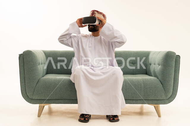 Looking at something, immersion and simulation of the imaginary world, enjoyment and entertainment through watching with virtual reality glasses, a portrait of an Arab Gulf Omani man wearing a dishdasha and turban sitting on a sofa wearing 3D glasses (VR), technological and technical development, white background.