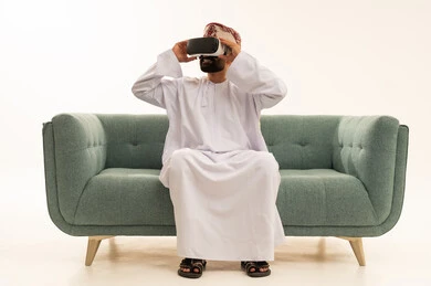 النظر الى شيء ما، الاندماج ومحاكاة العالم الخيالي، المتعة والتسلية بالمشاهدة عبر نظارة الواقع الافتراضي، بورتريه لرجل عربي خليجي عماني يرتدي الدشداشة و العمامة يجلس على الاريكة  يضع نظارات ثلاثية الأبعاد (الفي ار), التطور التقني والتكنولوجي، خلفية بيضاء