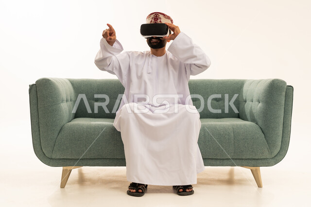 التفاعل والاندماج مع الواقع الافتراضي، قضاء وقت الفراغ بشكل ممتع، الاشارة بإصبع السبابة إلى شيء ما، استخدام الأجهزة التقنية الحديثة، رؤية المشاهد الخيالية، بورتريه لرجل عربي خليجي عماني يرتدي الدشداشة و العمامة يجلس على الاريكة يضع نظارات الفي آر، خلفية بيضاء