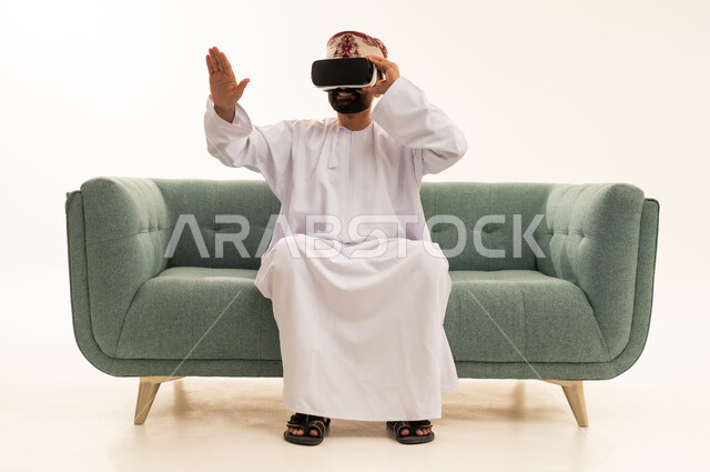 رفع كف اليد للأعلى بإيماءات الاندماج ومحاكاة العالم الخيالي، التطور التقني والتكنولوجي, المتعة بالمشاهدة عبر نظارة الواقع الافتراضي، بورتريه لرجل عربي خليجي عماني يرتدي الدشداشة و العمامة يجلس على الاريك ينظر من خلال نظارات ثلاثية الأبعاد (الفي ار)، خلفية بيضاء .