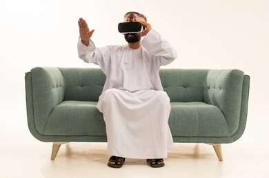 رفع كف اليد للأعلى بإيماءات الاندماج ومحاكاة العالم الخيالي، التطور التقني والتكنولوجي, المتعة بالمشاهدة عبر نظارة الواقع الافتراضي، بورتريه لرجل عربي خليجي عماني يرتدي الدشداشة و العمامة يجلس على الاريك ينظر من خلال نظارات ثلاثية الأبعاد (الفي ار)، خلفية بيضاء .