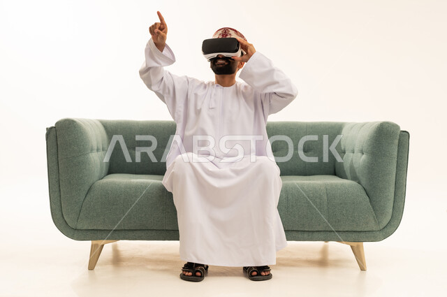 Spending free time in an enjoyable way, interacting and immersing oneself in virtual reality, pointing with the index finger at something, using modern technological devices, seeing imaginary scenes, a portrait of an Arab Gulf Omani man wearing a dishdasha and a turban sitting on a sofa wearing VR glasses, white background.