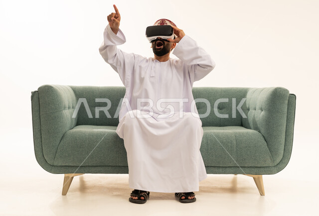 Gestures of astonishment and surprise, interaction and immersion with virtual reality, pointing with the index finger at something, using modern technological devices, seeing imaginary scenes, a portrait of an Arab Gulf Omani man wearing a dishdasha and a turban sitting on a sofa wearing VR glasses, white background.