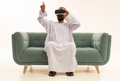 ايماءات الاندهاش و الاستغراب، التفاعل والاندماج مع الواقع الافتراضي، الاشارة بإصبع السبابة إلى شيء ما، استخدام الأجهزة التقنية الحديثة، رؤية المشاهد الخيالية، بورتريه لرجل عربي خليجي عماني يرتدي الدشداشة و العمامة يجلس على الاريكة يضع نظارات الفي آر، خلفية بيضاء