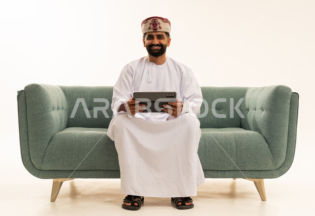 النظر للكاميرا بتعابير السرور، التطور التكنولوجي في مختلف المجالات, استخدام التقنيات الحديثة في العمل عن بعد، إنجاز الأعمال بسرعة وسهولة، بورتريه لرجل عربي خليجي عماني يرتدي الدشداشة و العمامة يجلس على الاريكة يحمل بيده جهاز التابلت ، تطبيقات وبرامج الأجهزة اللوحية ، خلفية بيضاء