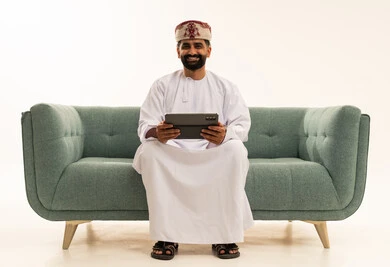 Looking at the camera with expressions of joy, technological advancement in various fields, using modern technologies for remote work, accomplishing tasks quickly and easily, a portrait of an Arab Gulf Omani man wearing a dishdasha and turban sitting on a sofa holding a tablet in his hand, applications and programs for tablets, white background.