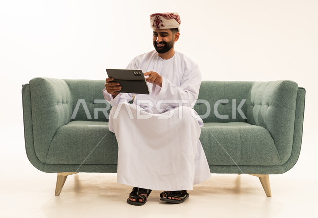 التطور التكنولوجي في مختلف المجالات, استخدام التقنيات الحديثة في العمل عن بعد، إنجاز الأعمال بسرعة وسهولة، بورتريه لرجل عربي خليجي عماني يرتدي الدشداشة و العمامة يجلس على الاريكة يحمل بيده جهاز التابلت ، النقر باصبع السبابة على الجهاز، تطبيقات وبرامج الأجهزة اللوحية ، خلفية بيضاء