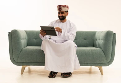 Technological development in various fields, the use of modern technologies in remote work, accomplishing tasks quickly and easily, a portrait of an Arab Gulf Omani man wearing a dishdasha and a turban sitting on a sofa holding a tablet in his hand, tapping with his index finger on the device, applications and programs for tablets, white background.