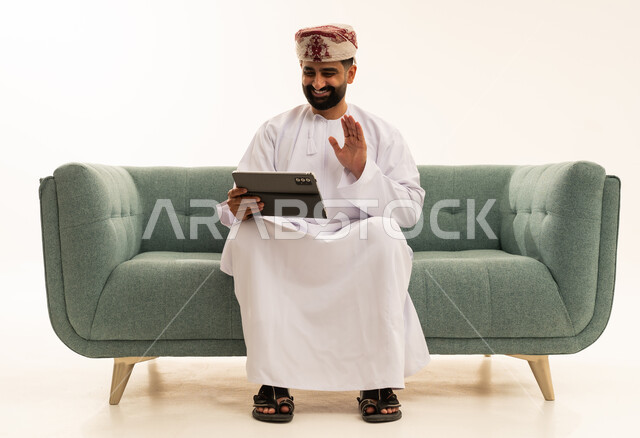 اجراء مكالمة فيديو، رفع كف اليد بتعابير السلام و التحية، استخدام التقنية الحديثه المتطورة في الحياة العملية، بورتريه لرجل عربي خليجي عماني يرتدي الدشداشة و العمامة يجلس على الاريكة يستخدم جهاز التابلت، خلفية بيضاء