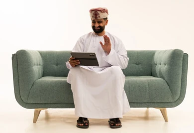 Making a video call, raising the palm with expressions of peace and greeting, using advanced modern technology in professional life, a portrait of an Arab Gulf Omani man wearing a dishdasha and a turban sitting on a sofa using a tablet, white background.