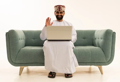 رفع كف اليد بتعابير السلام و التحية، اجراء مكالمة فيديو، العمل عن بعد، استخدام التقنية الحديثه المتطورة في الحياة العملية، بورتريه لرجل عربي خليجي عماني يرتدي الدشداشة و العمامة يجلس على الاريكة يقوم بالعمل على الحاسب المحمول، خلفية بيضاء