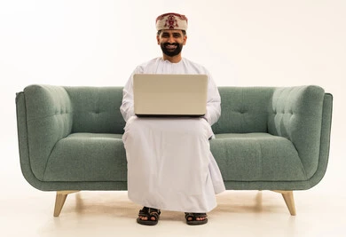 انجاز الاعمال والمهام عن بعد عبر الحاسوب المحمول، تصفح الانترنت ووسائل التواصل الاجتماعي،  بورتريه لرجل عربي خليجي عماني يرتدي الدشداشة و العمامة يجلس على الاريكة يقوم باستخدام الحاسب، خلفية بيضاء