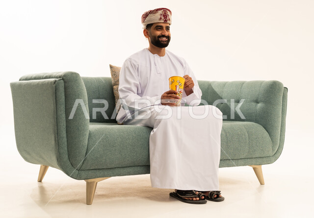 إيماءات الاندماج والسعادة والمتعة، مشاهدة الأفلام وإحضار السناكات اللذيذة، بورتريه لرجل عربي خليجي عماني يرتدي الدشداشة و العمامة يجلس على الاريكة يحمل بيده علبة الفوشار، قضاء أوقات ممتعة ومسلية، مفهوم التسلية والترفيه، خلفية بيضاء