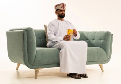 مشاهدة فيلم وإحضار السناكس الذيذ، بورتريه لرجل عربي خليجي عماني يرتدي الدشداشة و العمامة يجلس على الاريكة يحمل بيده علبة الفوشار، إيماءات الاندماج والسعادة والمتعة، قضاء أوقات ممتعة ومسلية، مفهوم التسلية والترفيه، خلفية بيضاء