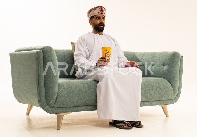ايماءات وجه تعبر عن الاندهاش، قضاء وقت ممتع برؤية فيلم سينمائي، الاستمتاع بتناول الفشار, بورتريه لرجل عربي خليجي عماني يرتدي الدشداشة و العمامة يجلس على الاريكة يقوم بتناول الفشار ومشاهدة الأفلام، خلفية بيضاء
