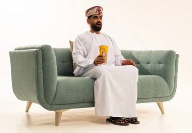 ايماءات وجه تعبر عن الاندهاش، قضاء وقت ممتع برؤية فيلم سينمائي، الاستمتاع بتناول الفشار, بورتريه لرجل عربي خليجي عماني يرتدي الدشداشة و العمامة يجلس على الاريكة يقوم بتناول الفشار ومشاهدة الأفلام، خلفية بيضاء