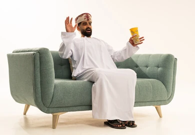 تعابير الخوف والذعر ،الاثارة والتشويق والاندماج، نشاط ترفيهي منزلي، مشاهدة فيلم و عرض مسرحي، بورتريه لرجل عربي خليجي عماني يرتدي الدشداشة و العمامة يجلس على الاريكة يحمل بيده دلو الفشار، تناول الوجبات الخفيفة والسناك، خلفية بيضاء