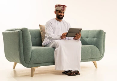 استخدام تطبيقات وبرامج الأجهزة اللوحية، تصفح مواقع التواصل الاجتماعي، الدردشة مع الأهل والأصدقاء، استعمال التكنولوجيا والتقنيات الحديثة،بورتريه لرجل عربي خليجي عماني يرتدي الدشداشة و العمامة يجلس على الاريكة يحمل بيده جهاز التابلت، خلفية بيضاء