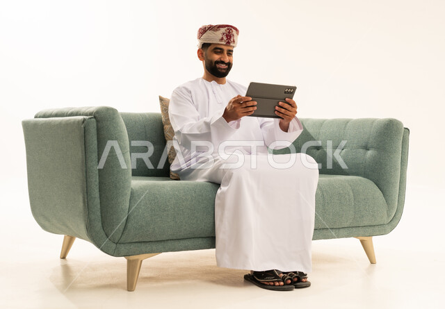 تصفح مواقع التواصل الاجتماعي، الدردشة مع الأهل والأصدقاء، استعمال التكنولوجيا والتقنيات الحديثة،بورتريه لرجل عربي خليجي عماني يرتدي الدشداشة و العمامة يجلس على الاريكة يحمل بيده جهاز التابلت، استخدام تطبيقات وبرامج الأجهزة اللوحية، خلفية بيضاء