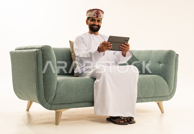 الدردشة مع الأهل والأصدقاء، استعمال التكنولوجيا والتقنيات الحديثة،بورتريه لرجل عربي خليجي عماني يرتدي الدشداشة و العمامة يجلس على الاريكة يحمل بيده جهاز التابلت، تصفح مواقع التواصل الاجتماعي، استخدام تطبيقات وبرامج الأجهزة اللوحية، خلفية بيضاء