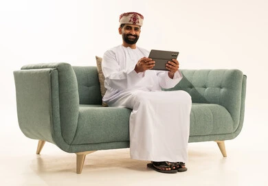 Chatting with family and friends, using technology and modern techniques, a portrait of an Arab Gulf Omani man wearing a dishdasha and a turban sitting on a sofa holding a tablet in his hand, browsing social media, using applications and programs for tablets, white background.