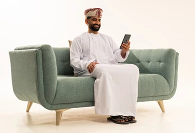 Browsing social media, using a modern technical device, communicating with family and friends, making a video call via mobile, a portrait of an Arab Gulf Omani man wearing a dishdasha and a turban sitting on a sofa and working on a mobile phone, white background.