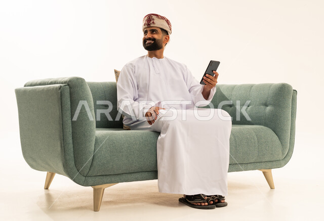 النظر الى شيء ما، تصفح مواقع التواصل الاجتماعي، استخدام جهاز تقني حديث، التواصل مع الأهل والاصدقاء، بورتريه لرجل عربي خليجي عماني يرتدي الدشداشة و العمامة يجلس على الاريكة  ويعمل على الهاتف المحمول، خلفية بيضاء