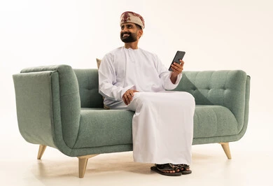 النظر الى شيء ما، تصفح مواقع التواصل الاجتماعي، استخدام جهاز تقني حديث، التواصل مع الأهل والاصدقاء، بورتريه لرجل عربي خليجي عماني يرتدي الدشداشة و العمامة يجلس على الاريكة  ويعمل على الهاتف المحمول، خلفية بيضاء